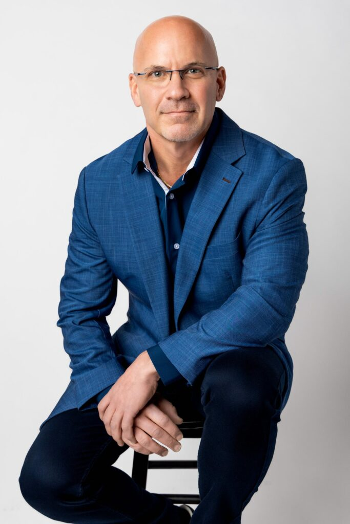 Bald man in a blue blazer sitting on a stool