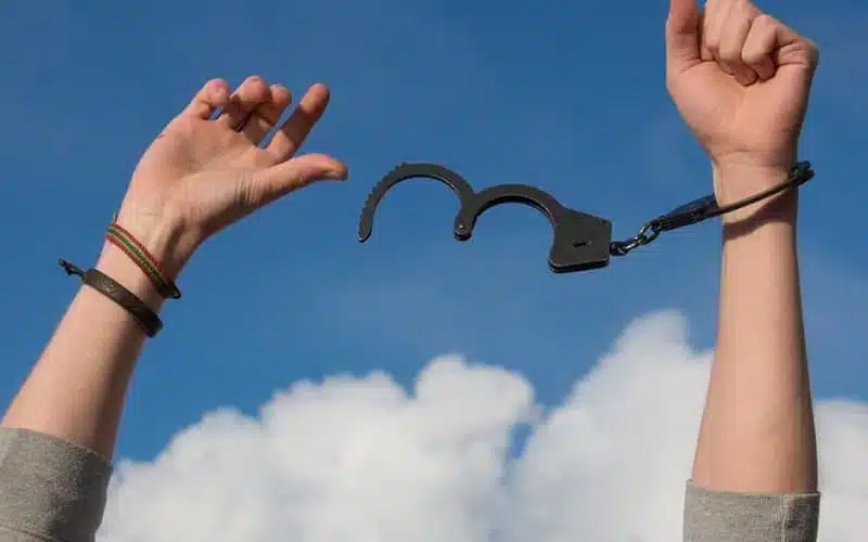 Two raised arms breaking apart a handcuff chain against a blue sky
