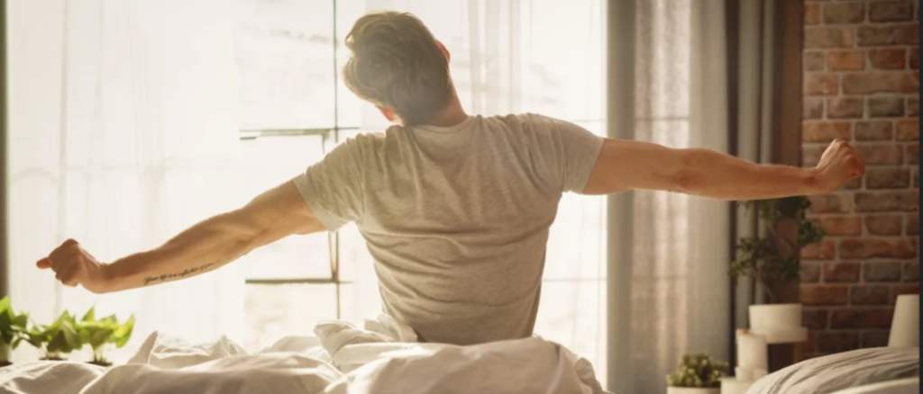 Man stretching his arms while sitting up in bed near a bright window